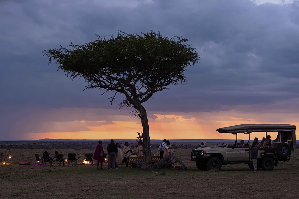 Guests gather beside a safari vehicle under a lone acacia tree as the sun sets beneath dramatic clouds over the African savanna