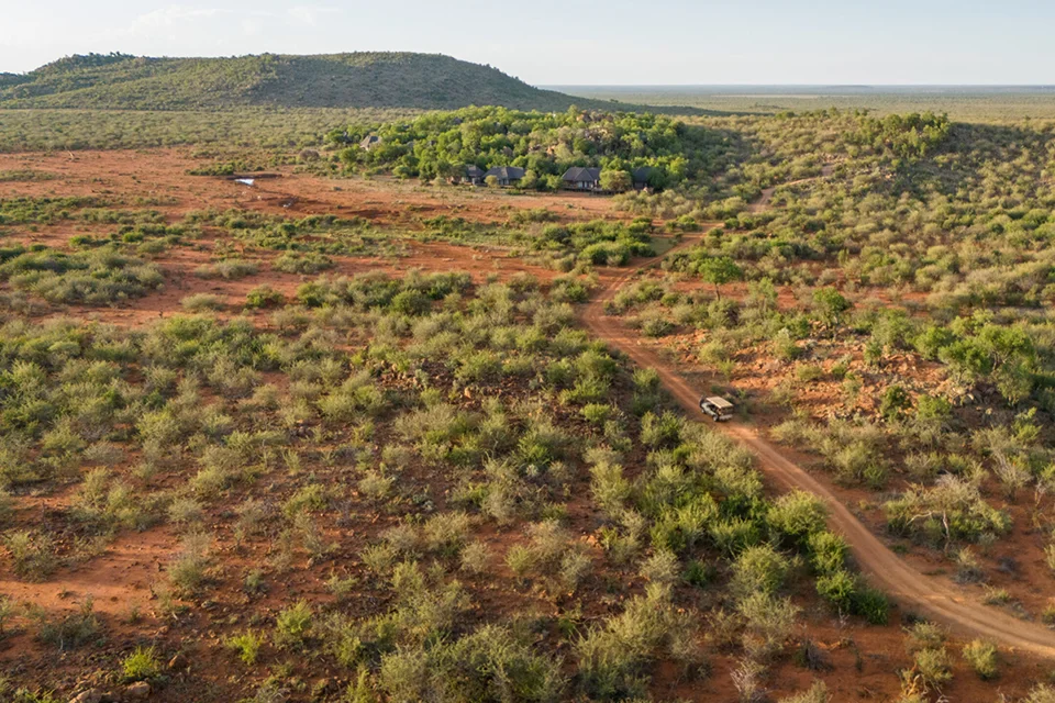 Aerial view of a safari vehicle travelling along a dirt road through a vast, green bushveld landscape, with a lodge nestled among the trees in the distance