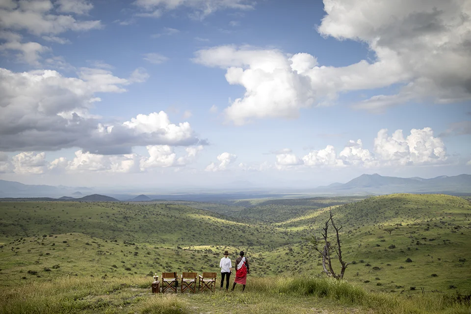 Two people stand beside safari chairs overlooking rolling green hills and expansive savanna beneath a dramatic, cloud-filled sky