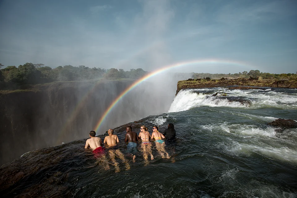 Visitors enjoying Devil's Pool on the edge of Victoria Falls beneath a vibrant rainbow