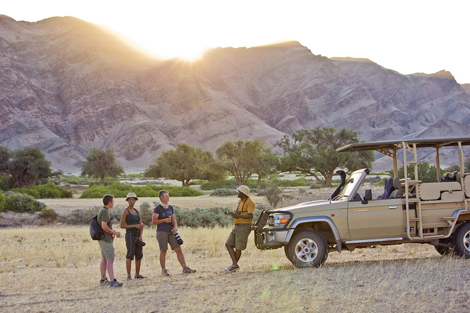 Guests chatting with a safari guide beside an open game-drive vehicle near Hoanib Valley Camp in northwest Namibia