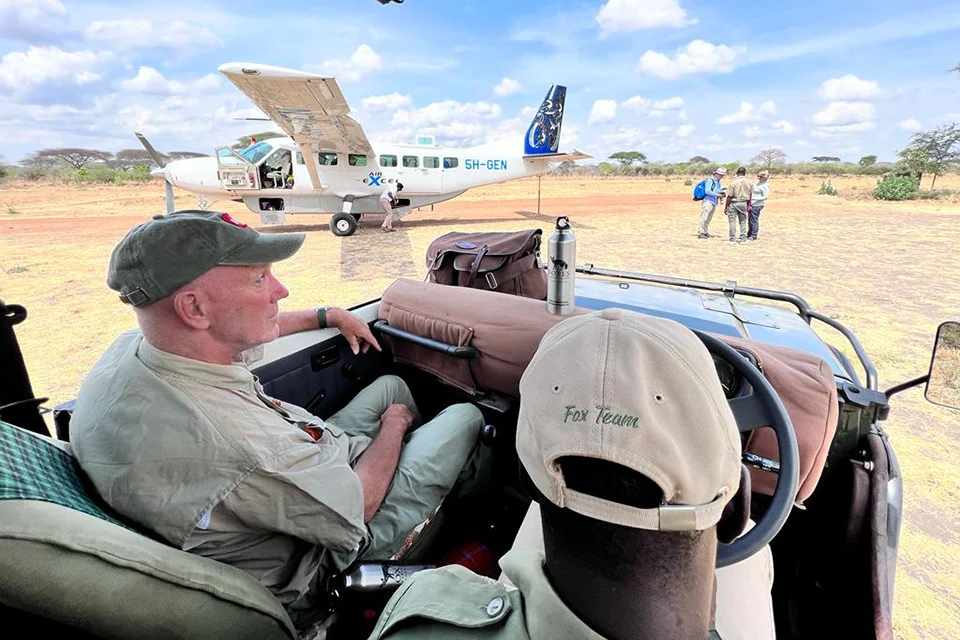 Guest seated in a wheelchair in an open safari vehicle beside an Air Excel aircraft at a remote airstrip in Tanzania