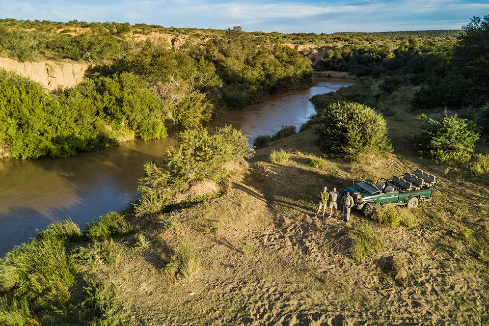 A safari vehicle and guests pause beside a winding river surrounded by lush vegetation in a remote African reserve