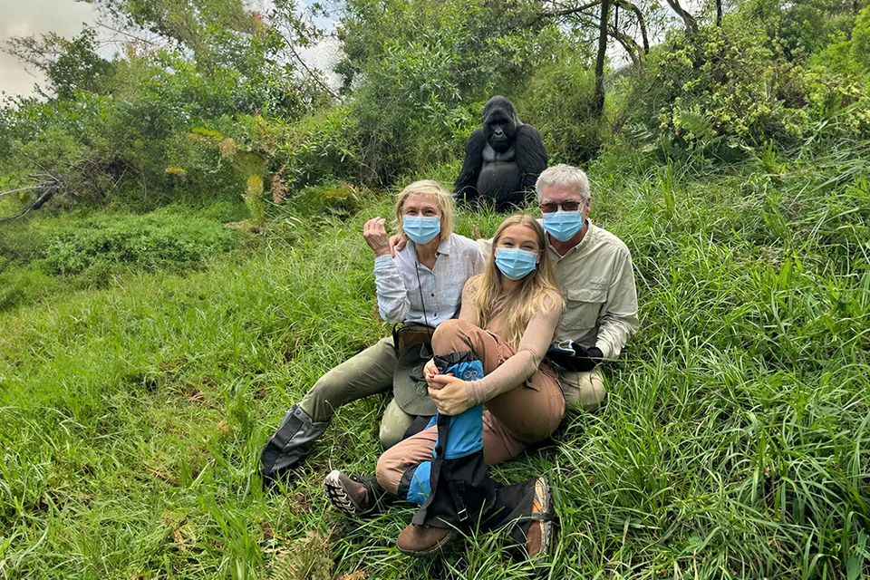 Three masked travellers sit together in lush green vegetation with a large gorilla resting on the hillside behind them