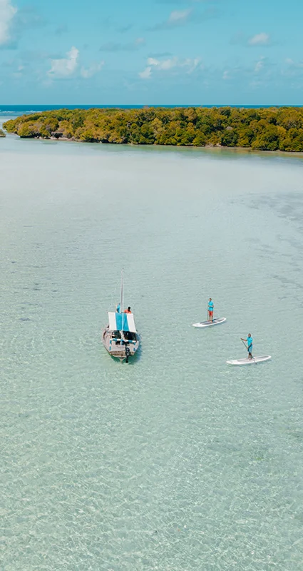Two guests stand-up paddleboarding beside a traditional boat in the shallow turquoise waters of Chole Mjini Lagoon, Mafia Island, Tanzania