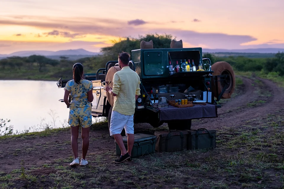 Couple enjoy a romantic sundowner beside a safari vehicle and drinks setup overlooking a waterhole at sunset during an African safari honeymoon
