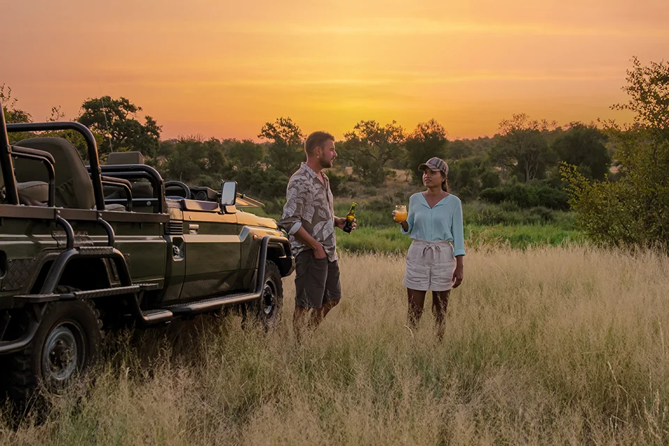 Couple enjoy sunset drinks beside a safari vehicle during a romantic game drive stop, surrounded by golden grassland under a glowing African sky