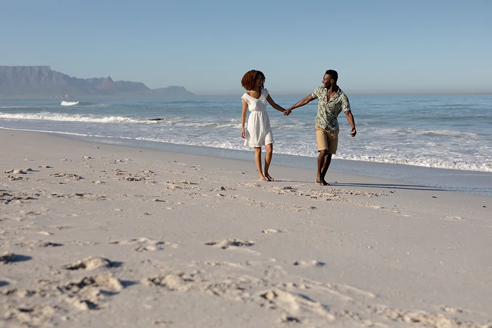 Couple walk hand in hand along a quiet sandy beach, enjoying a romantic coastal escape with ocean waves and distant mountains in the background
