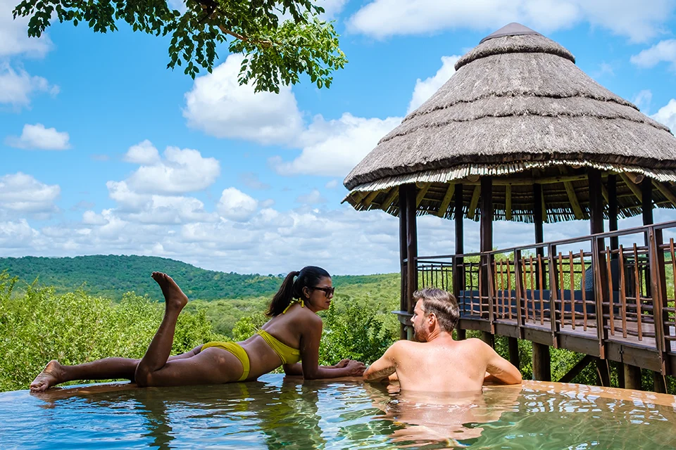 Couple relax together in a private plunge pool at a luxury safari lodge, overlooking rolling bushveld views and a traditional thatched pavilion.
