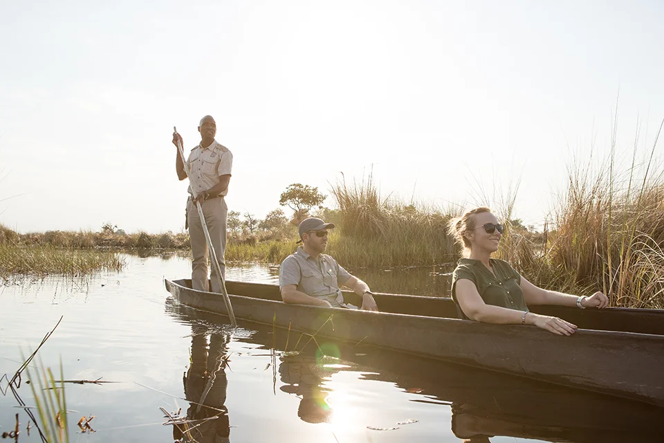 Couple enjoy a peaceful mokoro canoe safari through the Okavango Delta, guided along a sunlit waterway lined with reeds and wetland grasses