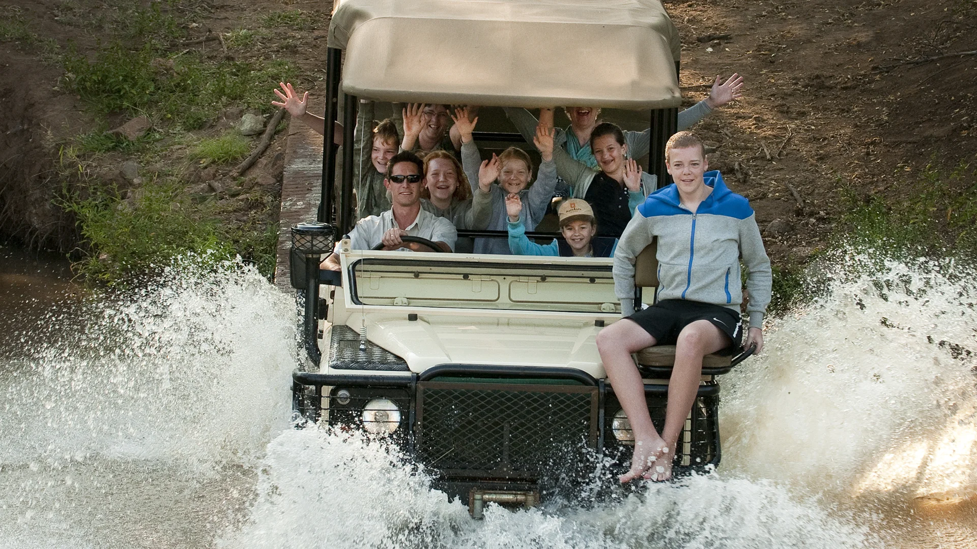 A safari vehicle drives through a shallow water crossing, sending up large splashes as a group of smiling adults and children wave toward the camera