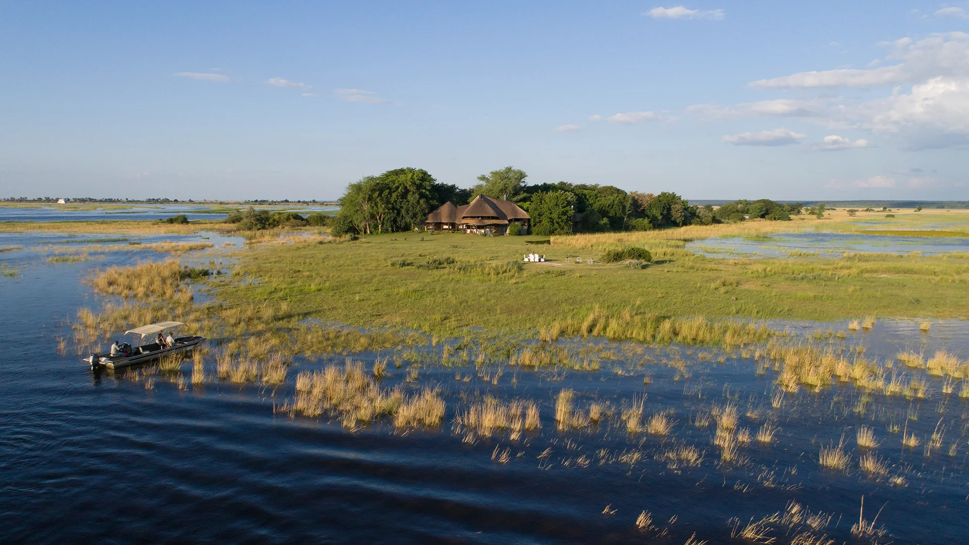 A safari lodge surrounded by green wetlands and shallow waterways, with a small motorboat crossing the water in the foreground