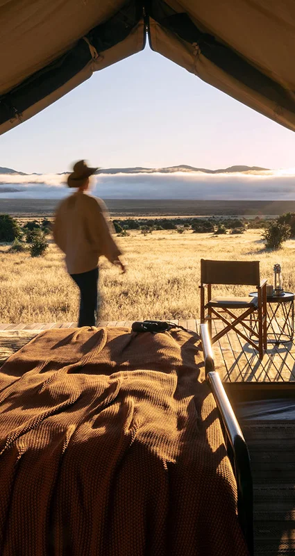 Luxury safari tent overlooking the Samara Karoo plains at sunrise, with a guest stepping onto the private deck and mountain views beyond