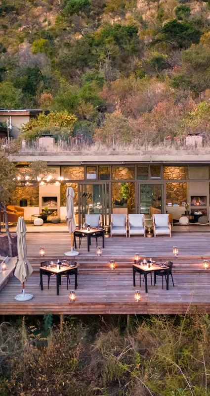 Outdoor dining deck at a safari lodge, surrounded by dense bushveld and set with candlelit tables