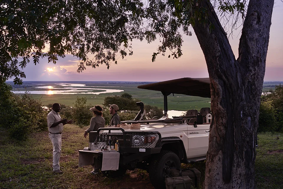 Safari guests enjoy drinks beside an open vehicle overlooking a vast wetland at sunset