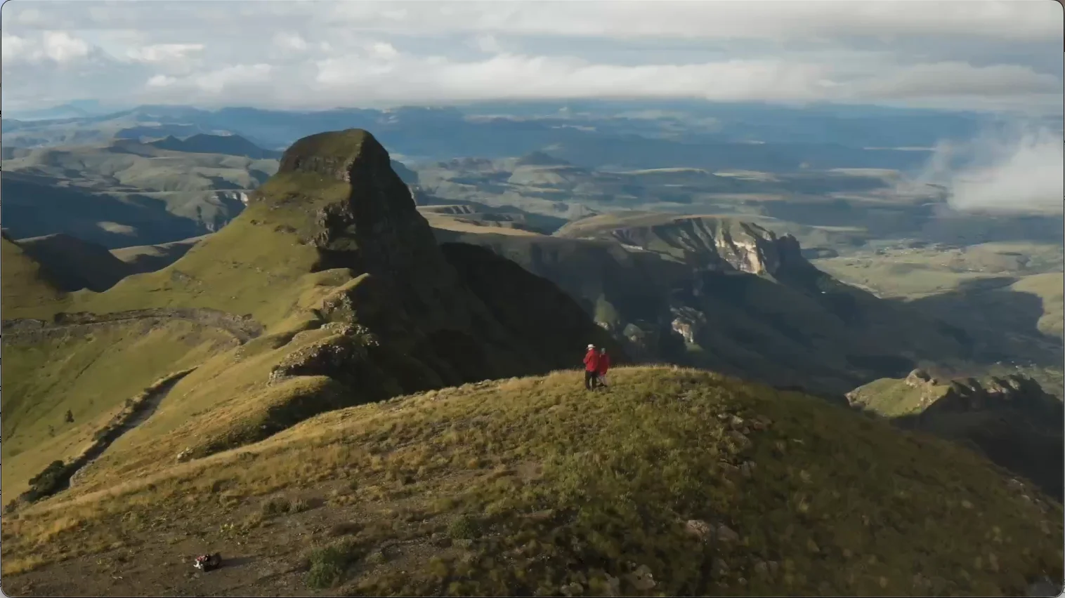 Two hikers standing on a grassy mountain ridge overlooking the dramatic peaks and valleys of the Drakensberg Mountains, KwaZulu-Natal