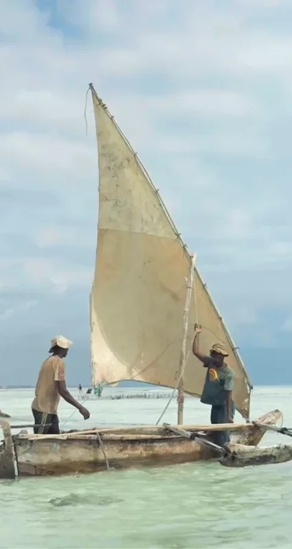 Two fishermen aboard a traditional wooden dhow with a raised sail in the shallow coastal waters near Saadani National Park, Tanzania