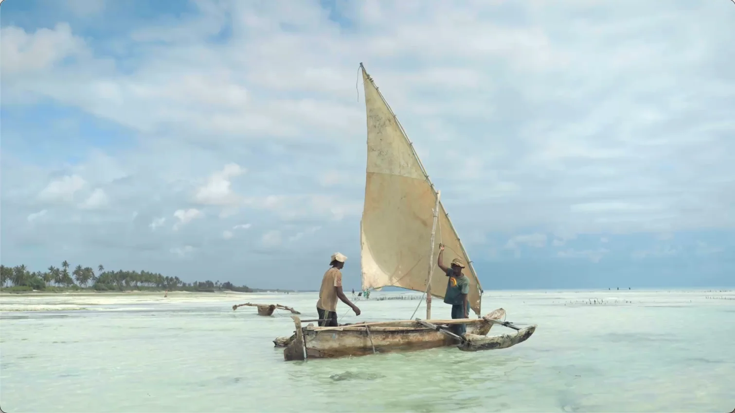 Two fishermen aboard a traditional wooden dhow with a raised sail in the shallow coastal waters near Saadani National Park, Tanzania