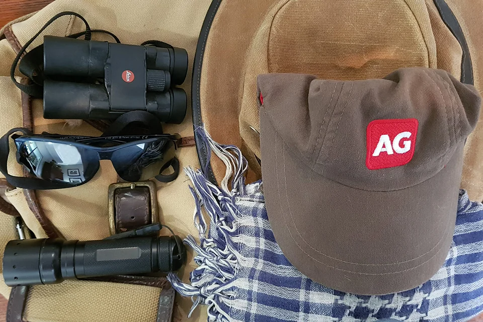 Safari essentials including binoculars, sunglasses, a sun hat, torch and cap laid out on a travel bag.