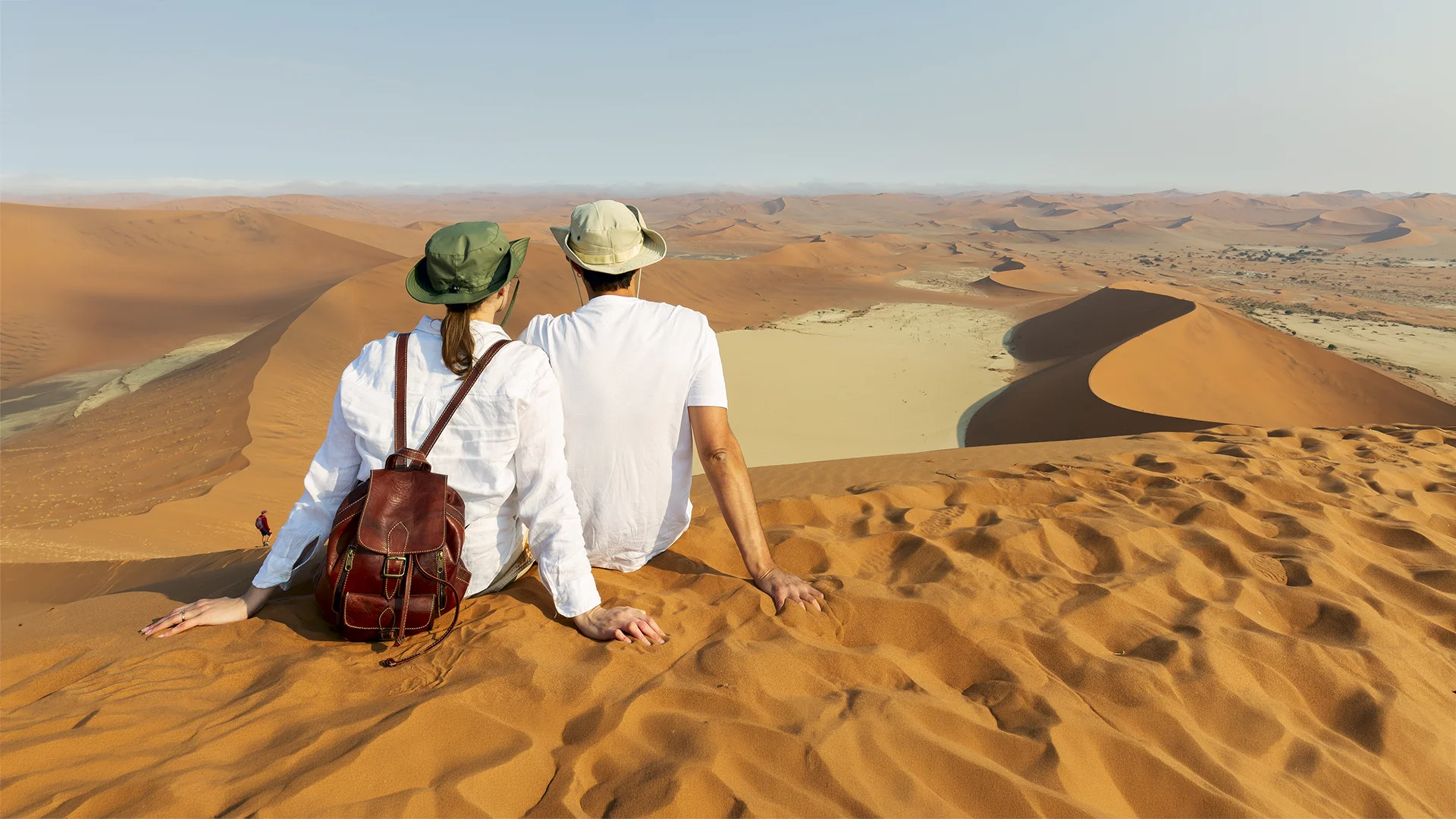 Two tourists sitting on a red sand dune overlooking the vast desert landscape of Sossusvlei in Namibia's Namib Desert