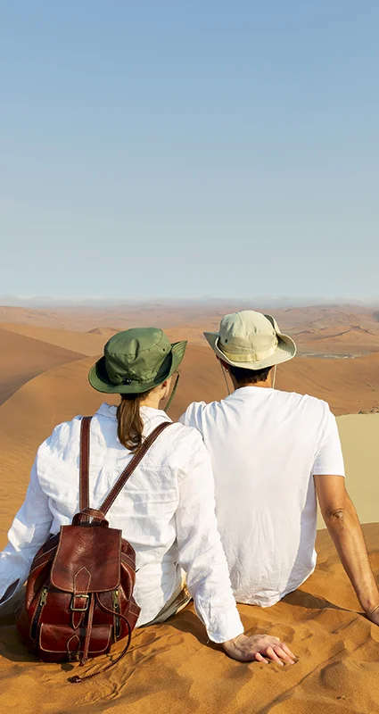 Two tourists sitting on a red sand dune overlooking the vast desert landscape of Sossusvlei in Namibia's Namib Desert
