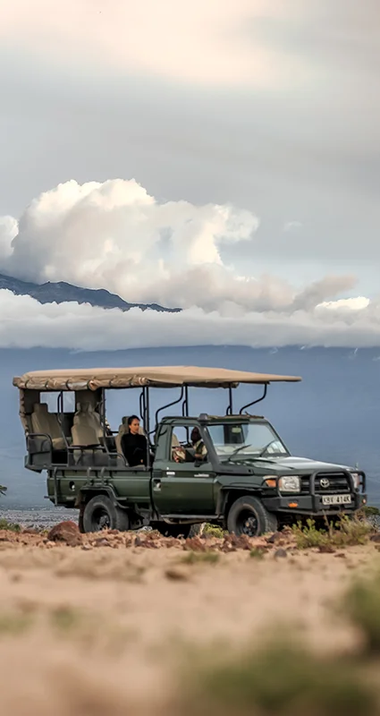 Open-air safari vehicle on a rocky plain with Mount Kilimanjaro rising through low clouds in the background