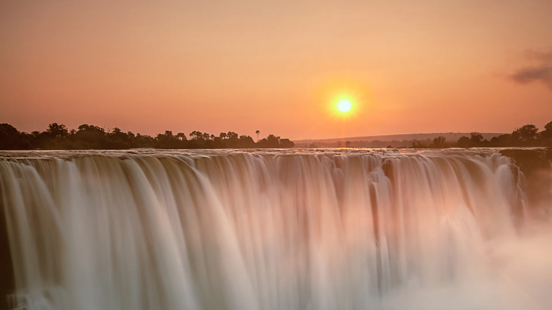 Victoria Falls at sunset, with the Zambezi River plunging over the cliff beneath a golden-orange sky