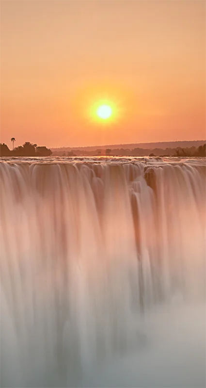 Victoria Falls at sunset, with the Zambezi River plunging over the cliff beneath a golden-orange sky