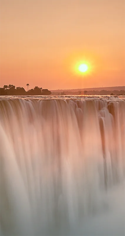 Victoria Falls at sunset, with golden light illuminating the cascading water and rising mist along the Zambezi River